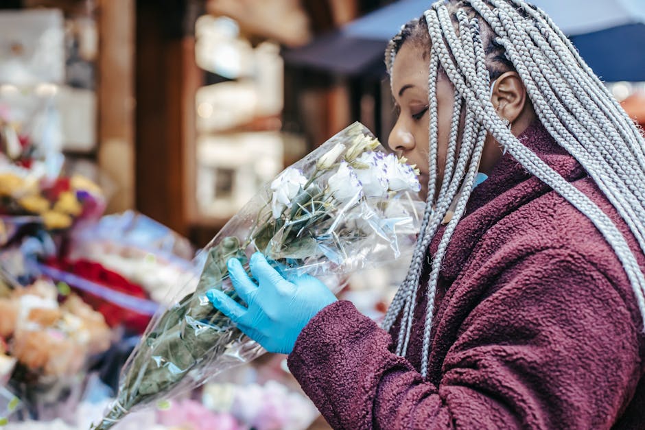 A woman with long, silver braided hair is holding a bouquet of fresh white roses wrapped in clear plastic at an outdoor flower stall. She is wearing a deep red, textured fleece jacket and bright blue