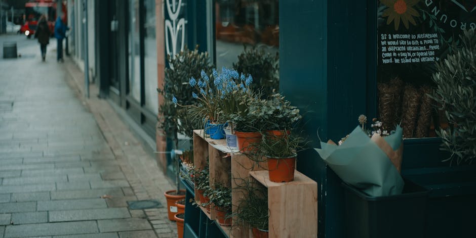 Close-up view of a small outdoor flower display outside a shop, featuring potted plants and floral arrangements in terracotta, blue, and white pots. The flowers include delicate blue blossoms and gree