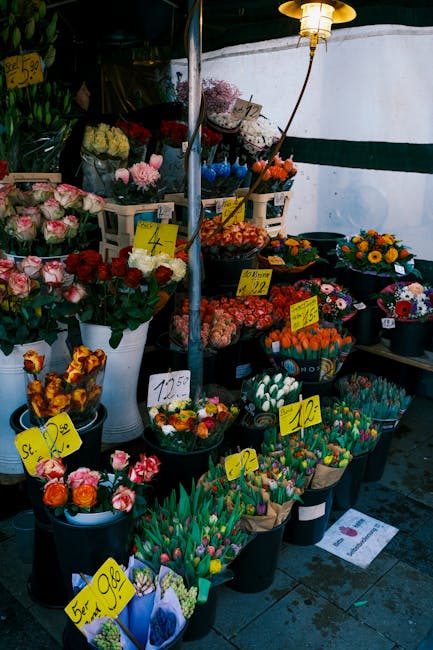 A vibrant display of fresh flowers arranged in black pots and white buckets at a flower stall, featuring a variety of blooms including pink and red roses, tulips in shades of pink, orange, and yellow,