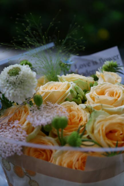 A close-up of a flower arrangement featuring pale peach roses with soft, velvety petals and creamy white blossoms, accented by small green buds and delicate white globe thistles. The flowers are arran