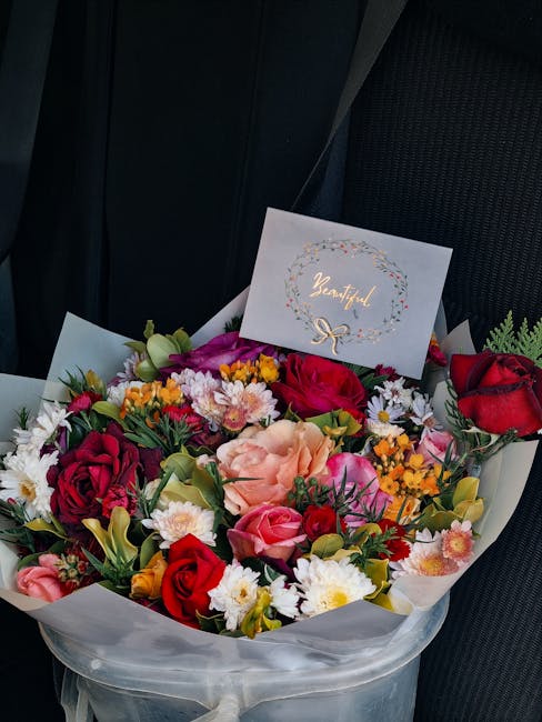 A vibrant bouquet of fresh flowers arranged in a round display, featuring red and pink roses, peach and purple carnations, white daisies, and small yellow blossoms, all wrapped in white tissue paper.