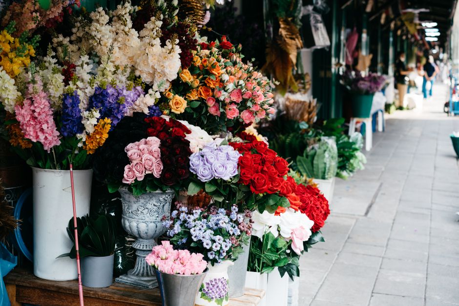 A storefront display of various fresh flower bouquets arranged in large white and ornate silver vases, featuring a vibrant mix of colors including pink, red, purple, white, and yellow. The flowers inc