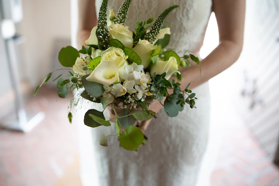 A person in a white lace dress holds a bouquet of fresh wedding flowers, featuring creamy white roses, delicate white orchid blossoms, and tall sprigs of green veronica. The arrangement includes vario
