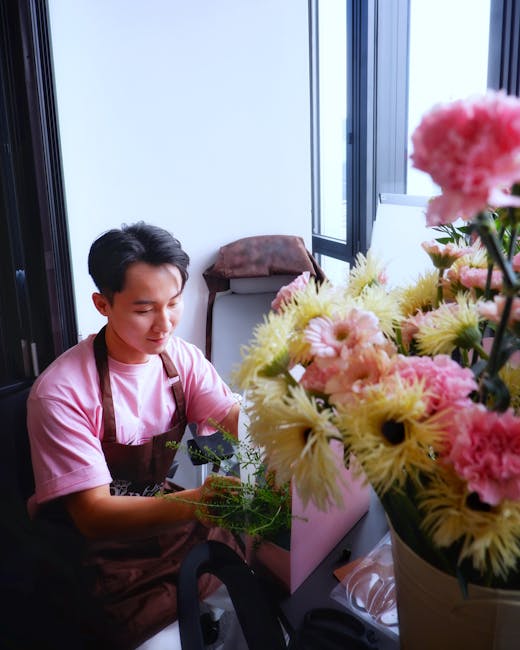 A young male florist with short dark hair, wearing a pink t-shirt and a brown apron, arranging a bouquet of fresh flowers inside a well-lit room with large windows. The bouquet includes pale yellow an