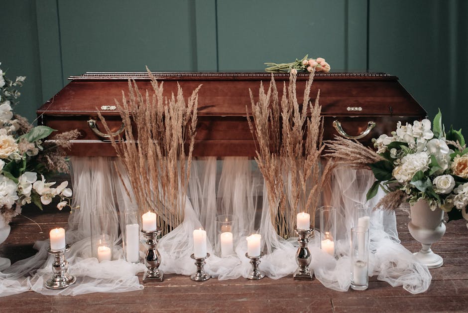 A funeral tribute featuring a closed wooden coffin adorned with a floral arrangement of white roses, pale pink carnations, and green foliage, flanked by two large white urns filled with similar white