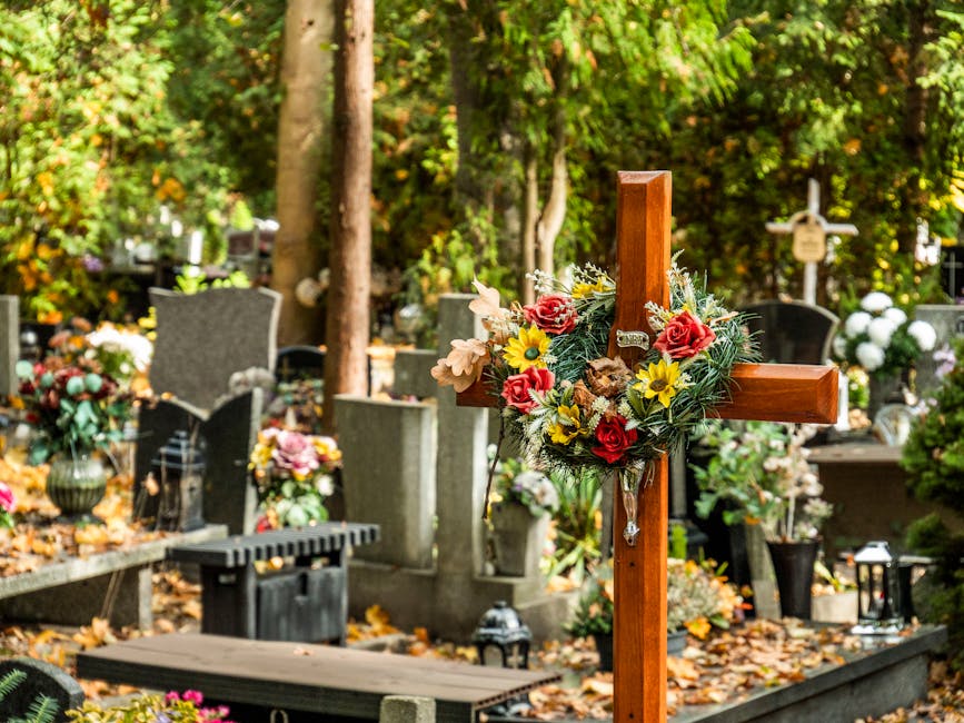 A wooden cross at a cemetery features a floral arrangement of fresh flowers including red roses, yellow daisies, and beige lilies, surrounded by green foliage. The arrangement is attached to the cross