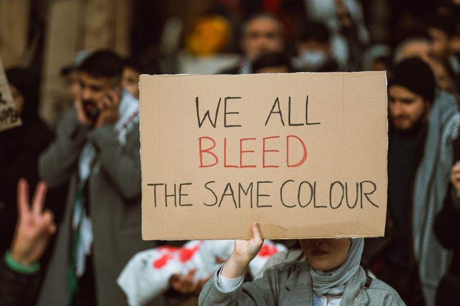 A group of protesters, some smiling and making peace signs, holding a large cardboard sign that reads 'We all bleed the same colour' in handwritten black and red letters. The protest appears to be ind